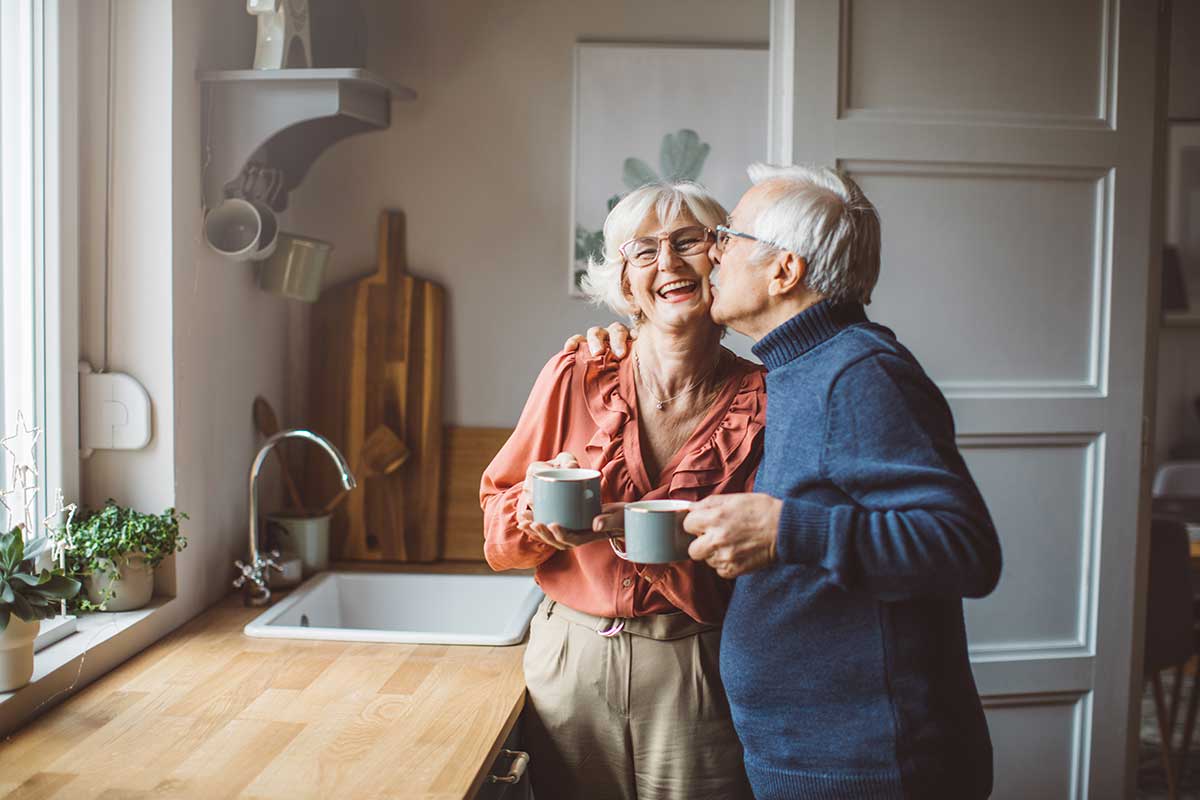 An older woman gets a kiss on the cheek from her husband while they hold mugs of coffee